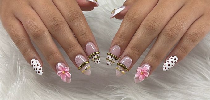 Almond nails on a sheer pink base mixing white nails with brown polka dots, raised pink flowers, white lace, and gold cuffs with tiny gold chains near the tips, over white fur.
