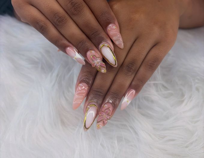 Long almond nails on a sheer pink base with raised pink flowers, thin gold outlines, small pearls and white swirls.